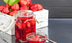 Słoik pełen truskawek w zalewie, obok miseczka z owocami. W tle drewniana skrzynka z dojrzałymi truskawkami. To domowy ocet truskawkowy.