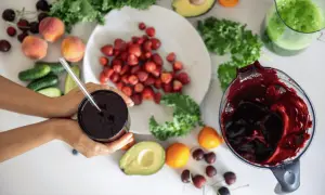 Ręce trzymają kubek z ciemnym sokiem, obok składniki na smoothie: truskawki, brzoskwinie, awokado, jarmuż, ogórki i wiśnie. Pomyśl o tym jak o naturalnym sposobie, jak rozszerzyć naczynia krwionośne.