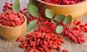 Suszone jagody goji na co? W drewnianych miseczkach i na stole leżą czerwone jagody goji, obok gałązka z świeżymi owocami.
