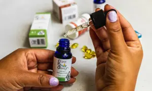 Dłoń trzymająca pipetkę z kroplą olejku CBD nad małą, niebieską buteleczką. W tle widać opakowania produktów z CBD, sugerujące jego działanie.