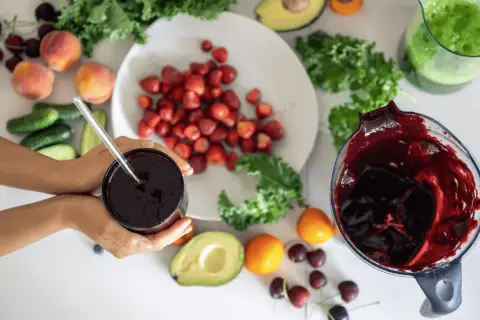 Ręce trzymają kubek z ciemnym sokiem, obok składniki na smoothie: truskawki, brzoskwinie, awokado, jarmuż, ogórki i wiśnie. Pomyśl o tym jak o naturalnym sposobie, jak rozszerzyć naczynia krwionośne.