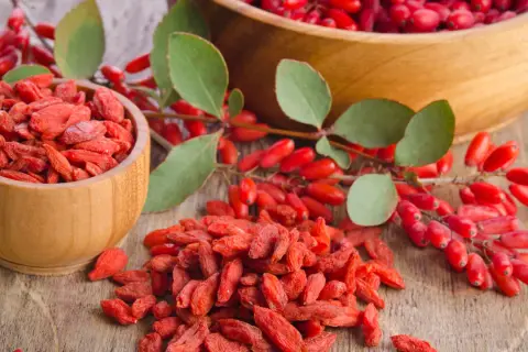 Suszone jagody goji na co? W drewnianych miseczkach i na stole leżą czerwone jagody goji, obok gałązka z świeżymi owocami.