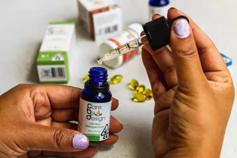 Dłoń trzymająca pipetkę z kroplą olejku CBD nad małą, niebieską buteleczką. W tle widać opakowania produktów z CBD, sugerujące jego działanie.