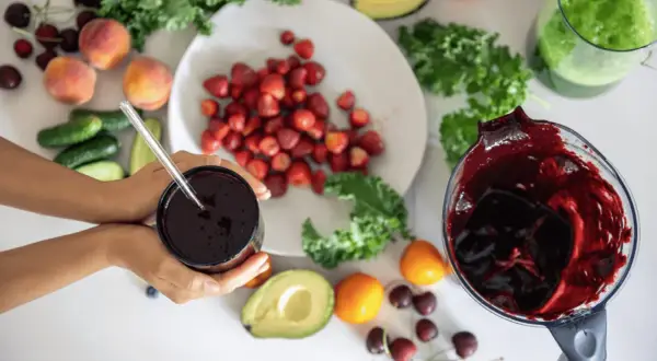 Ręce trzymają kubek z ciemnym sokiem, obok składniki na smoothie: truskawki, brzoskwinie, awokado, jarmuż, ogórki i wiśnie. Pomyśl o tym jak o naturalnym sposobie, jak rozszerzyć naczynia krwionośne.