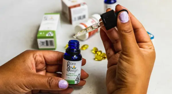 Dłoń trzymająca pipetkę z kroplą olejku CBD nad małą, niebieską buteleczką. W tle widać opakowania produktów z CBD, sugerujące jego działanie.
