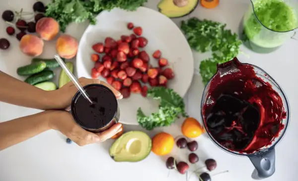 Ręce trzymają kubek z ciemnym sokiem, obok składniki na smoothie: truskawki, brzoskwinie, awokado, jarmuż, ogórki i wiśnie. Pomyśl o tym jak o naturalnym sposobie, jak rozszerzyć naczynia krwionośne.