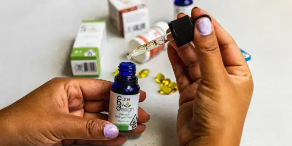 Dłoń trzymająca pipetkę z kroplą olejku CBD nad małą, niebieską buteleczką. W tle widać opakowania produktów z CBD, sugerujące jego działanie.